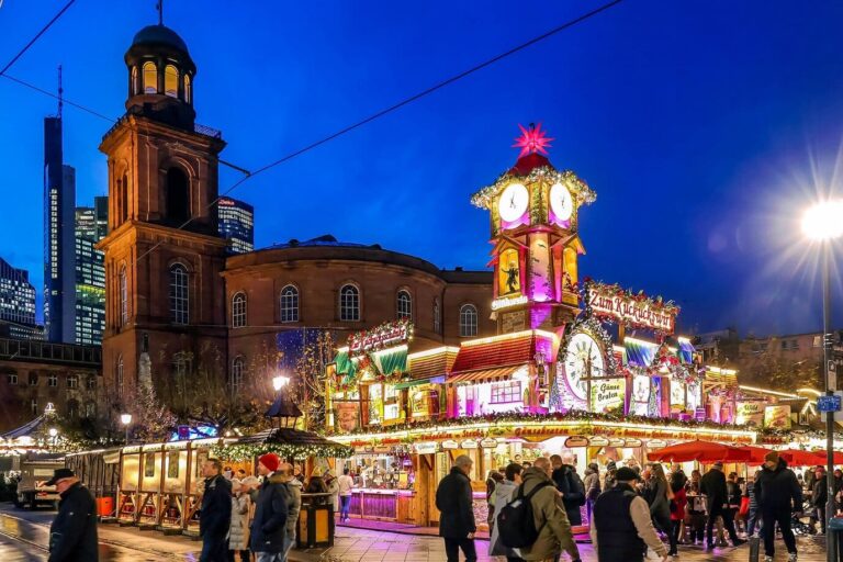Chalets illuminés et kiosque à horloge du marché de Noël devant l’église Saint-Paul, sur la Paulsplatz, avec les gratte-ciel de Francfort au loin, à la tombée de la nuit