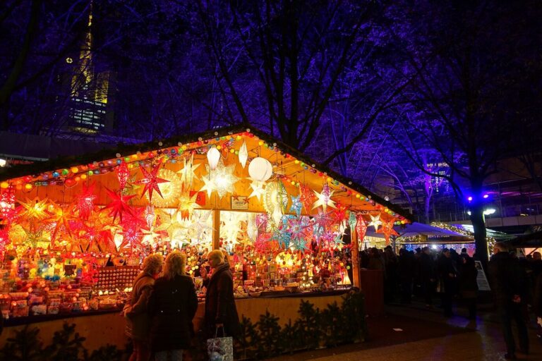 Chalet du marché de Noël couvert d’étoiles en papier lumineuses rouges et orangées, sous les arbres éclairés, la nuit