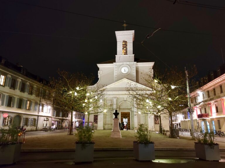 L’église Sainte-Croix de Carouge éclairée la nuit, devant sa place bordée d’arbres illuminés