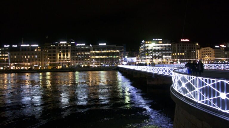 Passerelle bordée de lumières blanches au-dessus du Rhône, face aux quais illuminés de Genève
