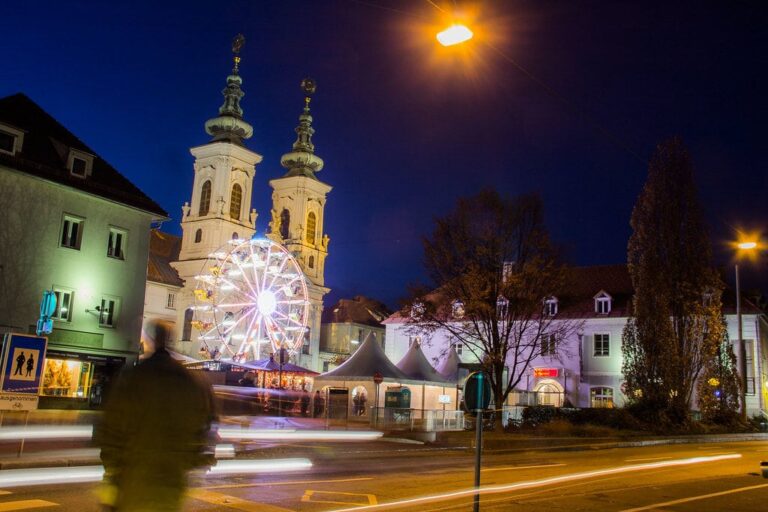 Grande roue illuminée et chalets du marché de Noël de la Mariahilferplatz, au pied des deux clochers baroques de l’église Mariahilf de Graz, à l’heure bleue