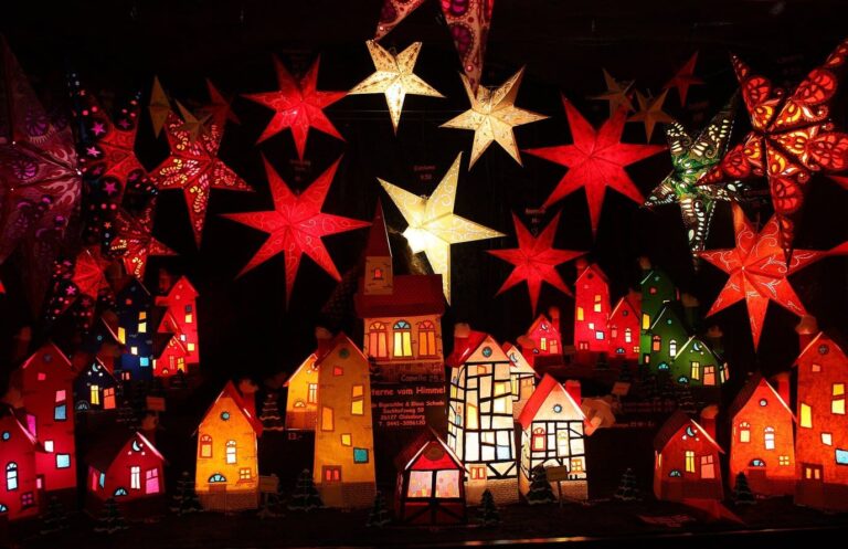 Des étoiles en papier rouges et dorées suspendues au-dessus de petites maisons-lanternes illuminées, sur un stand du marché