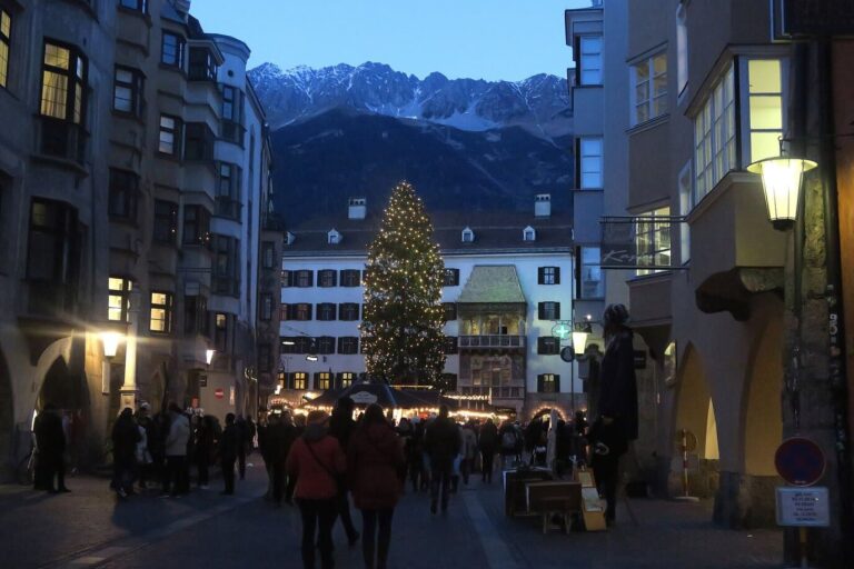 Rue de la vieille ville d’Innsbruck menant au sapin illuminé et au Petit Toit d’or, avec la chaîne enneigée de la Nordkette en arrière-plan, au crépuscule