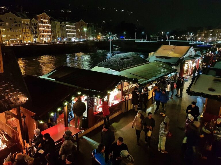 Allées de chalets éclairés du marché de la Marktplatz vues d’en haut, au bord de l’Inn, avec les façades colorées de l’autre rive, la nuit