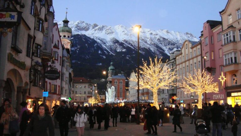 Arbres lumineux et foule sur la Maria-Theresien-Strasse d’Innsbruck à la tombée du jour, avec les sommets enneigés de la Nordkette au fond