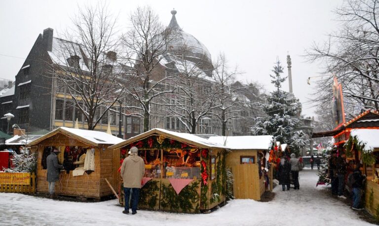 Allée de chalets en bois sous la neige au village de Noël de Liège, devant un bâtiment à coupole