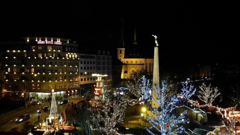 Le monument de la Gëlle Fra entouré des arbres illuminés du marché de Noël, la nuit, avec la cathédrale en arrière-plan