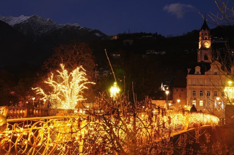 Le pont de la Poste de Merano couvert de guirlandes lumineuses, un arbre illuminé et le clocher de la ville sous les montagnes, à la nuit tombée