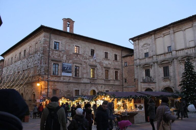 Chalets illuminés et promeneurs sur la piazza Grande à la tombée du jour, devant le palais communal et le palais Tarugi
