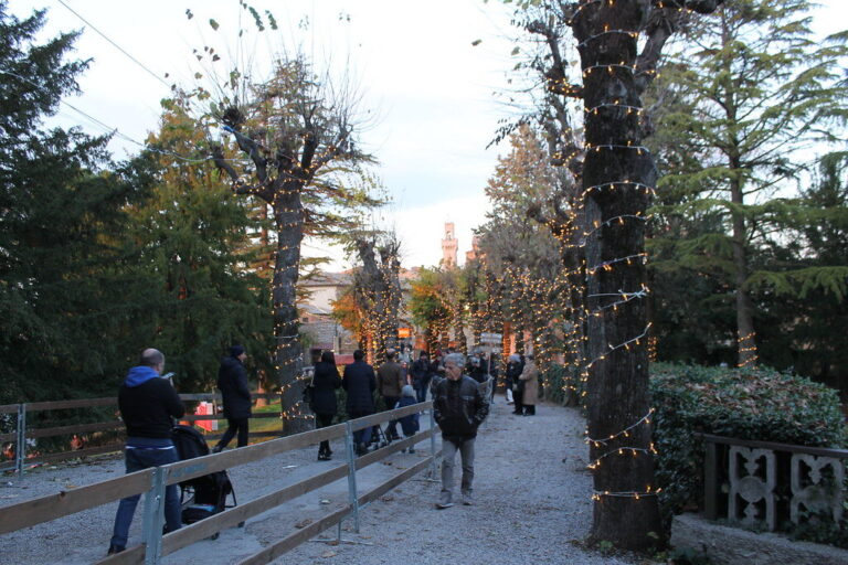 Allée bordée d’arbres enroulés de guirlandes lumineuses, avec des promeneurs et une tour du bourg à l’arrière-plan