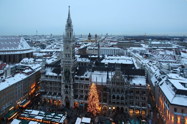 Vue plongeante sur la Marienplatz enneigée à l’heure bleue : Nouvel Hôtel de Ville, sapin illuminé et chalets du marché de Noël, avec les toits blancs de Munich jusqu’à l’horizon