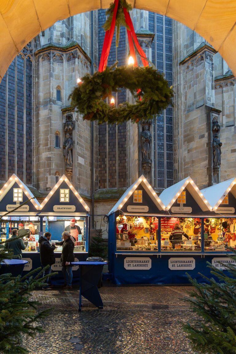 Une couronne de l’Avent suspendue sous une arcade en pierre, ouvrant sur les chalets bleus illuminés au pied de l’église Saint-Lambert