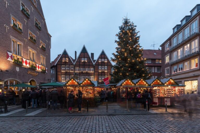 Le sapin illuminé et les chalets du village de Noël du Kiepenkerl, au Spiekerhof, devant des maisons à pignons, à l’heure bleue