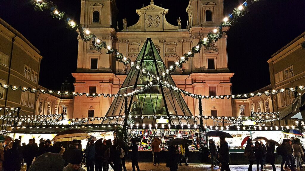 La façade illuminée de la cathédrale de Salzbourg derrière la pyramide de verre et les guirlandes du marché de Noël de la Domplatz, la nuit