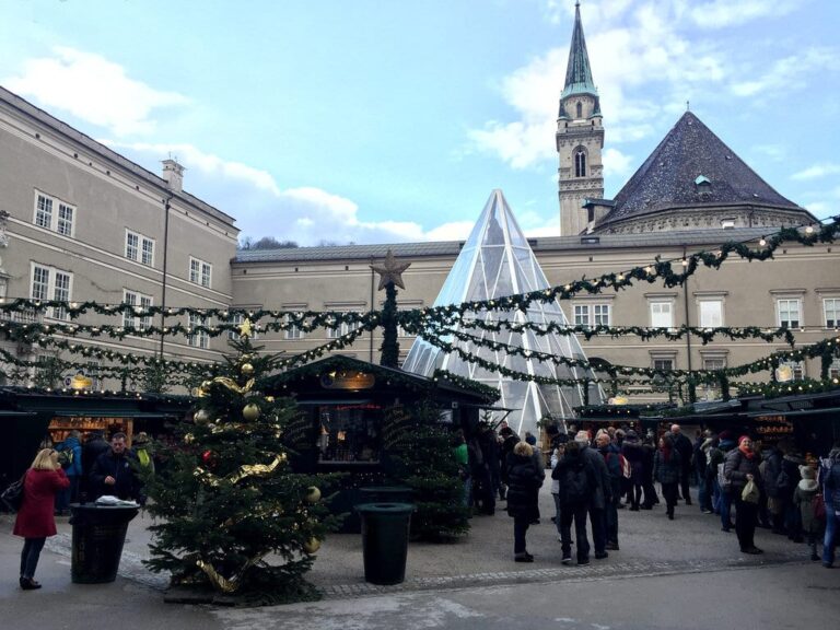 Chalets, sapin décoré et pyramide de verre protégeant la colonne de la Vierge sur la Domplatz de Salzbourg, avec le clocher de l’église des Franciscains en arrière-plan