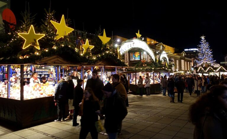 Allée de chalets illuminés sous des étoiles dorées, avec l’arche lumineuse et le grand sapin de la Schlossplatz de Stuttgart en fond, la nuit