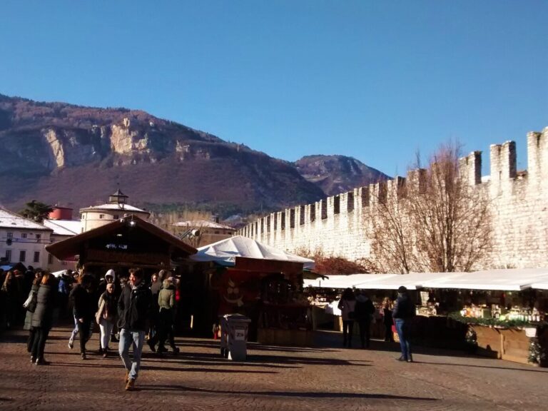 Chalets du marché de Noël au pied des remparts crénelés de la piazza Fiera, la montagne en arrière-plan, par un jour d’hiver ensoleillé