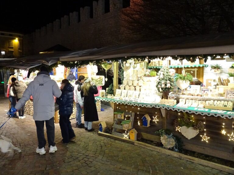 Visiteurs devant un chalet garni d’objets de bois et de décorations, le long des remparts éclairés