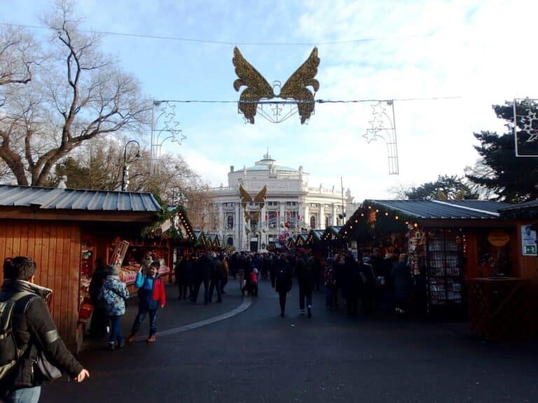 Allée de chalets du marché du Rathausplatz sous une guirlande en forme d’ange, avec le Burgtheater en arrière-plan