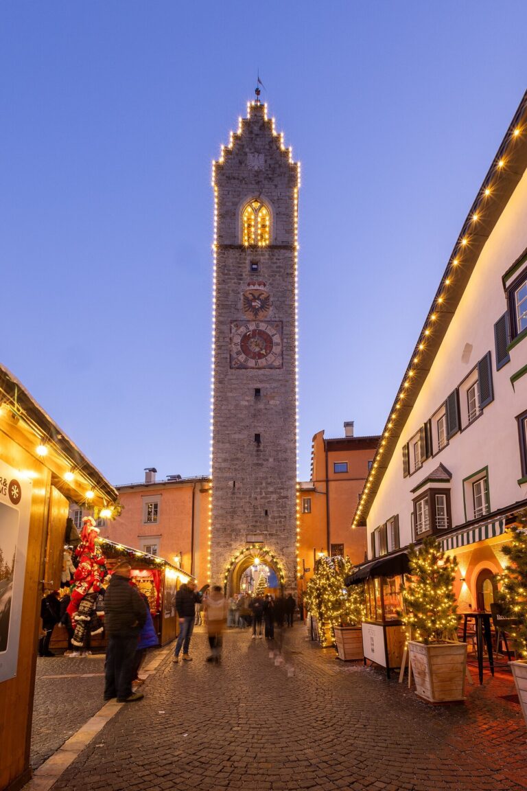La Torre delle Dodici illuminée au bout de la rue principale de Vipiteno, bordée de façades décorées, à l’heure bleue