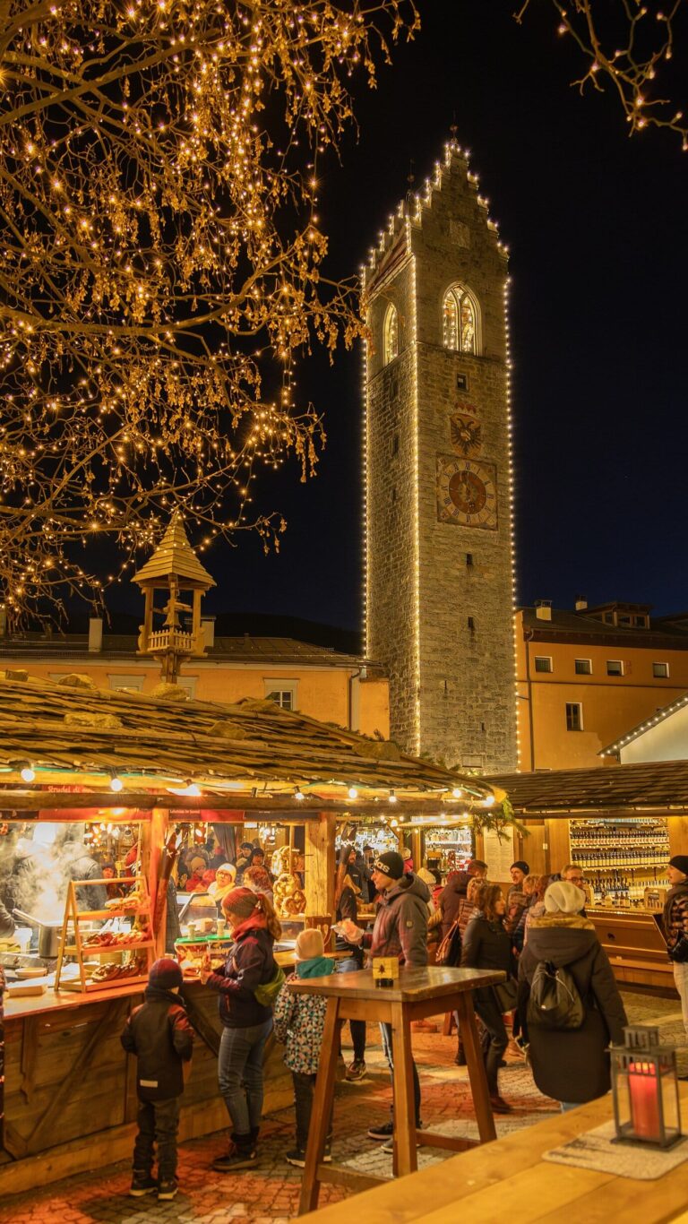 Chalet du marché de Noël sous des branches couvertes de lumières, la Torre delle Dodici éclairée en arrière-plan