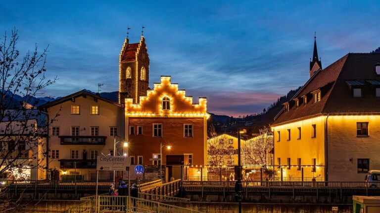 Maisons à pignons soulignés de guirlandes lumineuses et tour de Vipiteno, à l’heure bleue, sous les montagnes