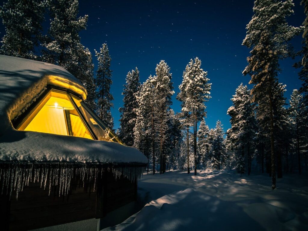 Une cabine à toit vitré éclairée de l’intérieur, chargée de neige, entre des pins givrés sous un ciel de nuit