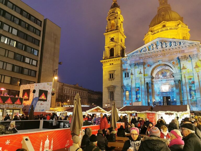Les deux tours de la basilique Saint-Étienne illuminées derrière la patinoire rouge et les chalets du marché de l’Avent, à la tombée de la nuit