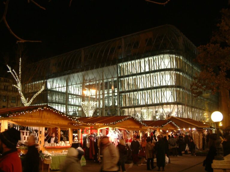 Les chalets illuminés du marché de Noël de la place Vörösmarty à Budapest, de nuit, devant l’immeuble de verre qui borde la place