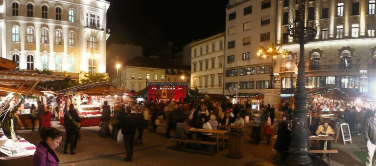 Des chalets, des tables et des passants sous les lampadaires du marché de Noël de la place Vörösmarty, de nuit, avec les façades éclairées au fond