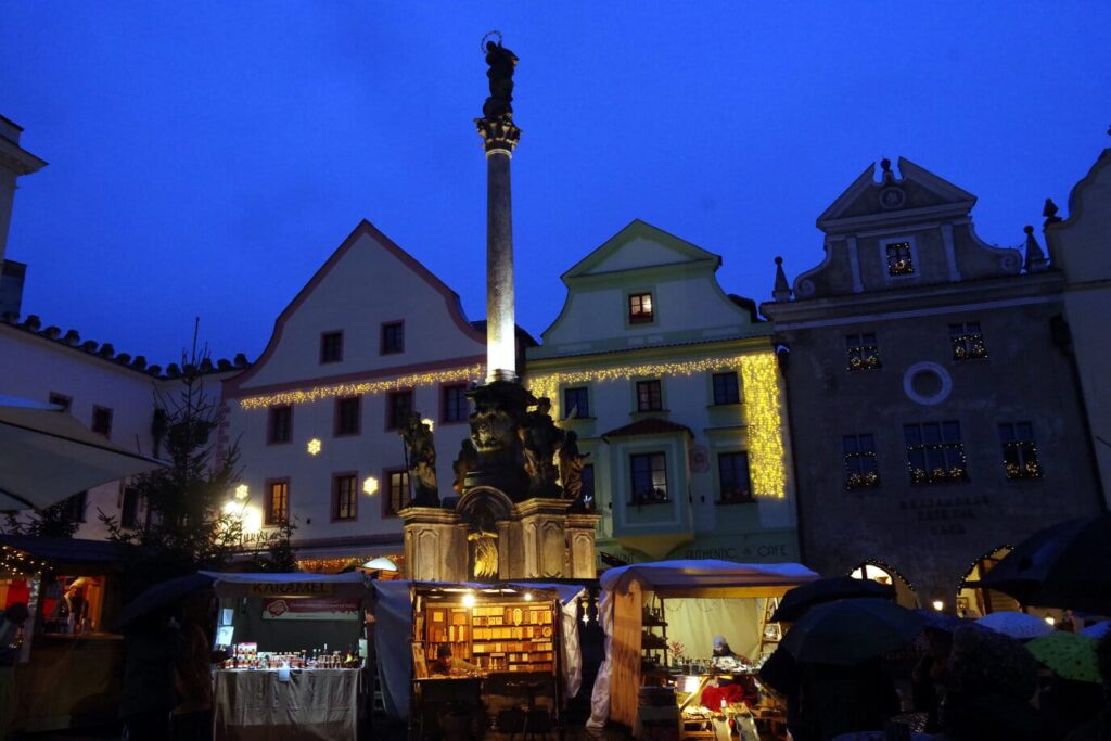 Les chalets illuminés du marché de Noël de la place Svornosti à Český Krumlov, au pied de la colonne de la peste et des maisons à pignons, à l’heure bleue