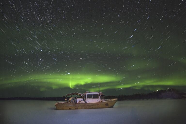 Les traînées d’étoiles et une aurore verte au-dessus du lac Inari gelé, avec un bateau échoué sur la glace