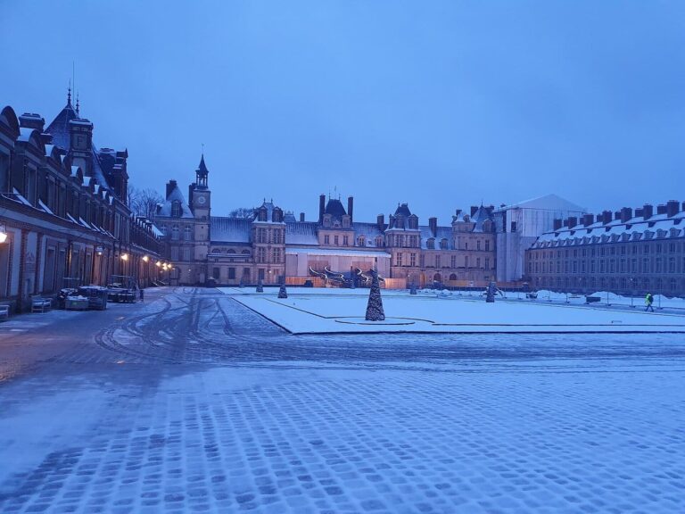 La cour du Cheval Blanc enneigée à l’heure bleue, avec le château au fond et un sapin décoratif au milieu de l’esplanade