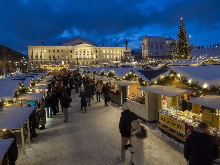 Une allée de chalets enneigés et décorés de branches de sapin au marché de Noël Tuomaan Markkinat, à la tombée de la nuit, devant les bâtiments de la place du Sénat