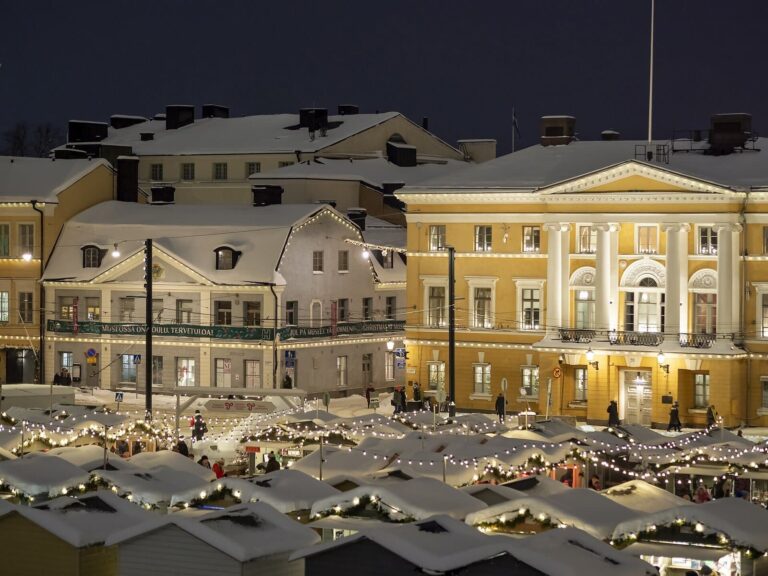 Les toits enneigés et guirlandés des chalets du marché de Noël de la place du Sénat, devant les façades jaunes éclairées de la place, la nuit