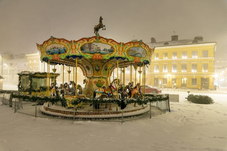 Le manège ancien du marché de Noël d’Helsinki sous une neige dense, guirlandes de verdure autour de la piste, façades jaunes éclairées derrière