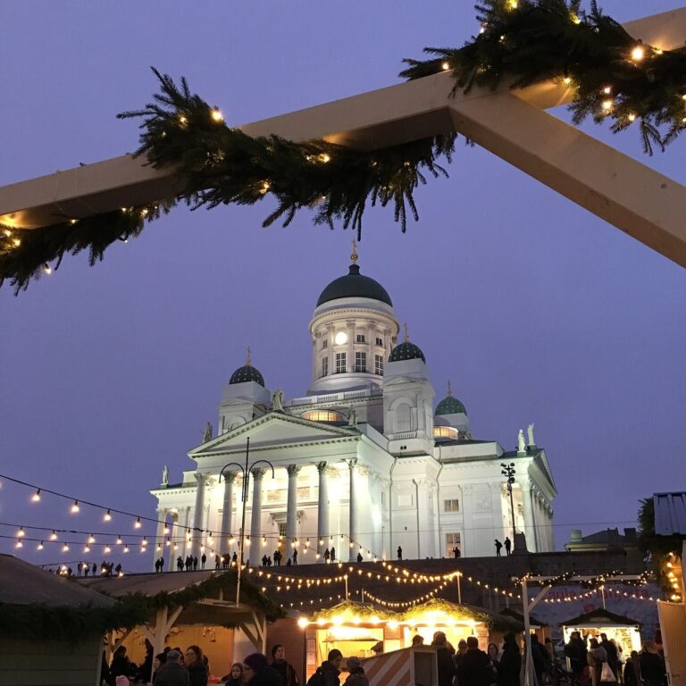 La cathédrale d’Helsinki illuminée au crépuscule, encadrée par une arche de branches de sapin et de lumières à l’entrée du marché de Noël