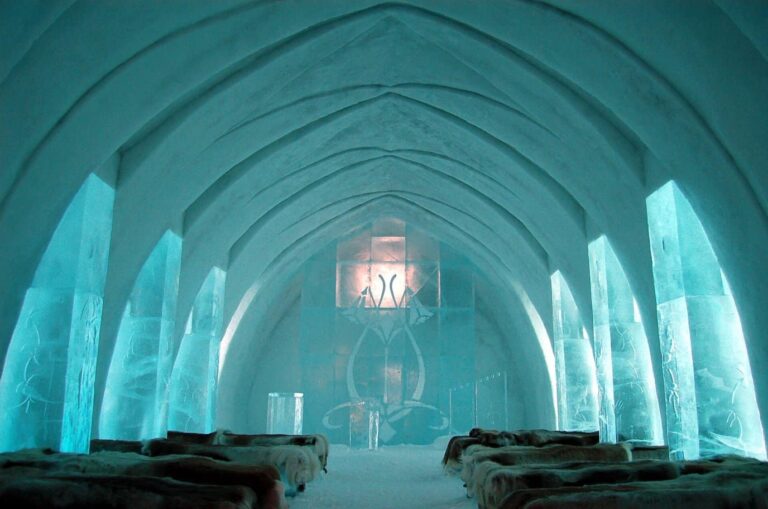 La nef voûtée de l’église de glace de l’Icehotel, éclairée en turquoise, avec ses bancs recouverts de peaux