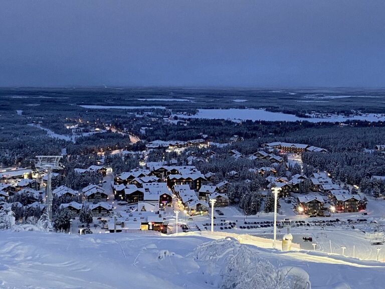 Le village de Sirkka et la station de Levi sous la neige à la tombée du jour, vus depuis les pentes du fell, avec les lumières des chalets et la forêt jusqu’à l’horizon