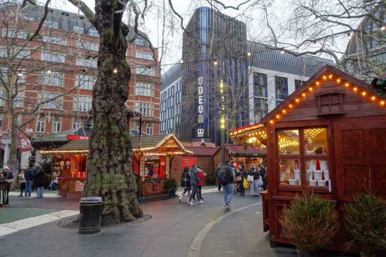 Une allée de chalets en bois du marché de Noël de Leicester Square, sous les grands arbres de la place, avec les immeubles de Londres en arrière-plan