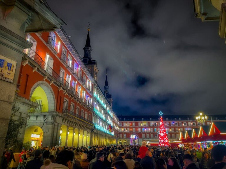 La Plaza Mayor de Madrid de nuit pendant le marché de Noël, ses façades éclairées en couleurs, un grand sapin lumineux rouge et la foule
