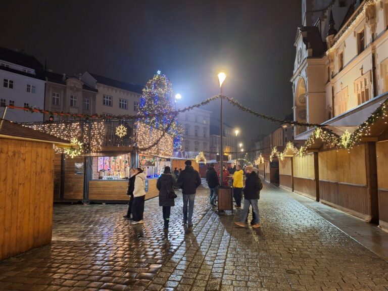 Chalets du marché de Noël d’Olomouc le long de l’hôtel de ville, guirlandes et sapin bleu en fond, un soir de pluie