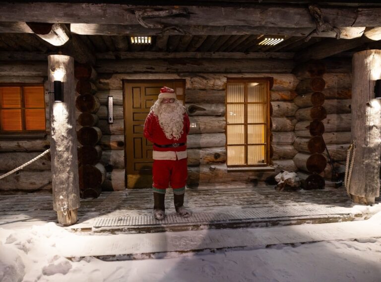 Le Père Noël debout devant la porte d’un chalet de rondins enneigé aux fenêtres éclairées, à Saariselkä, en Laponie finlandaise, en décembre 2024