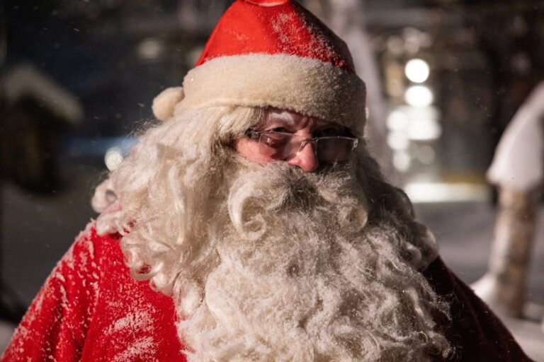 Le visage du Père Noël en gros plan, barbe blanche, lunettes et bonnet rouge saupoudré de neige, à Saariselkä, en décembre 2024
