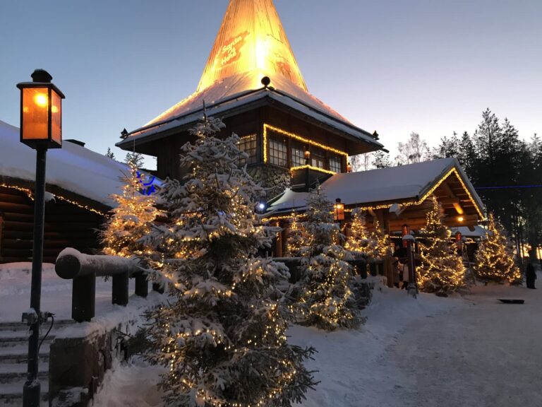 Le bâtiment principal du Santa Claus Village au crépuscule, toits enneigés et sapins couverts de guirlandes allumées, à Rovaniemi