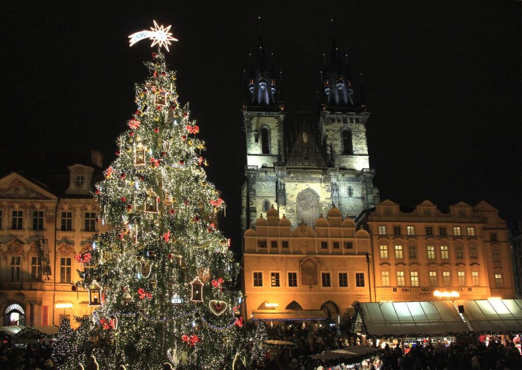 Le grand sapin illuminé de la place de la Vieille-Ville de Prague devant l’église Notre-Dame de Týn éclairée, avec les chalets du marché de Noël en contrebas, de nuit