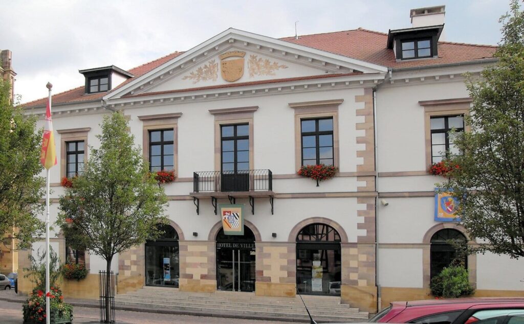 L’hôtel de ville de Rouffach, façade claire au fronton sculpté et balcon fleuri, encadrée de deux arbres