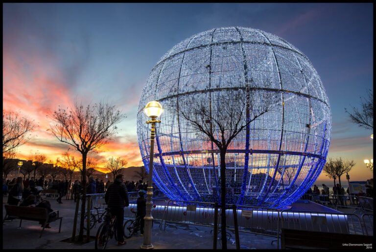 Une boule lumineuse géante blanche et bleue sur le front de mer de Saint-Sébastien, au coucher du soleil, entre des arbres nus et des réverbères