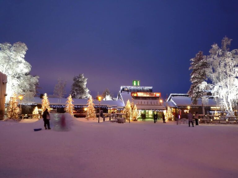 La place du Santa Claus Village le soir, avec les sapins illuminés et les bâtiments éclairés sous un ciel bleu nuit