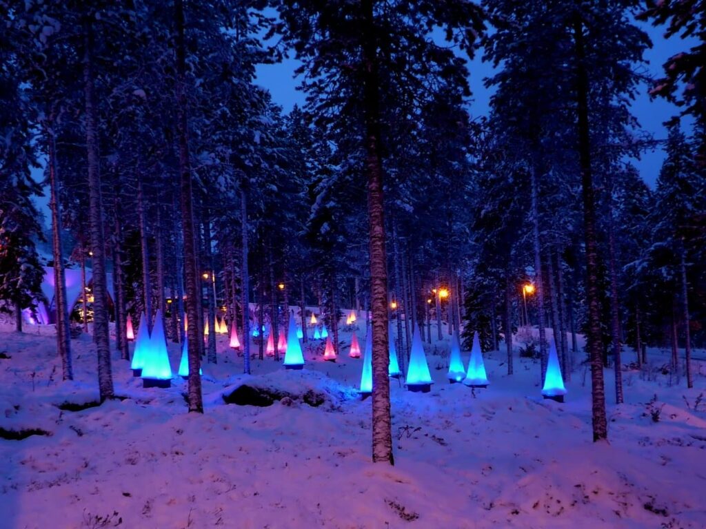 Des cônes lumineux bleus et roses plantés dans la neige entre les pins, à l’entrée de SantaPark, à la tombée de la nuit